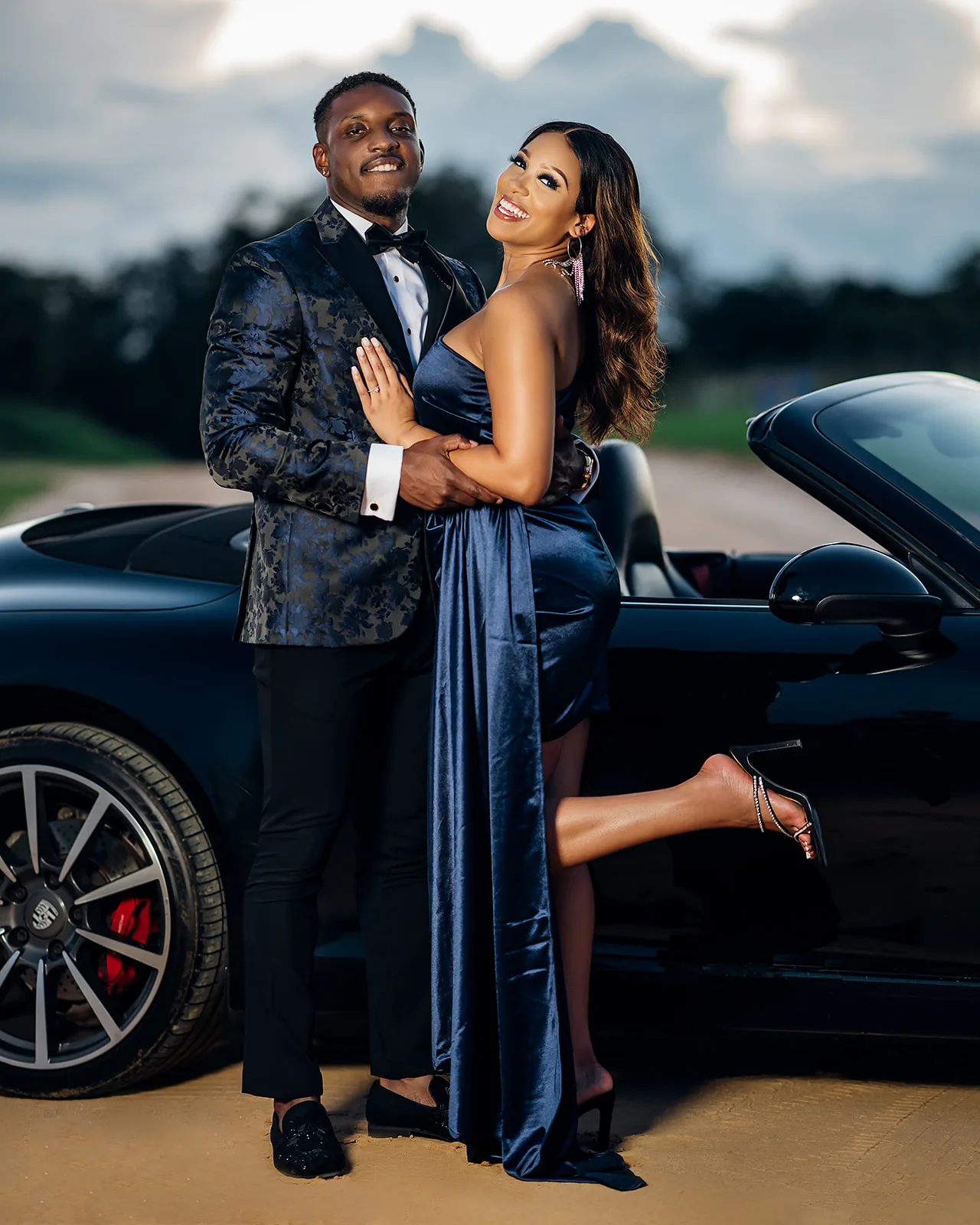 Glamorous couple posing beside black Porsche at golden hour