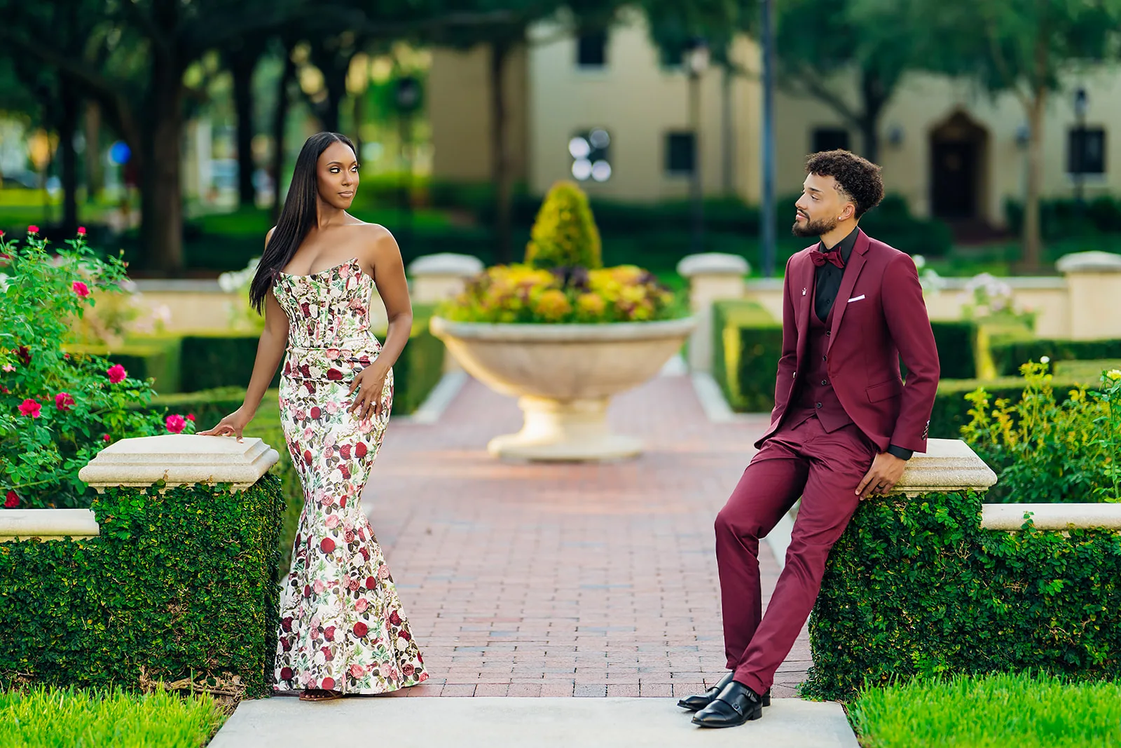 Couple in floral gown and burgundy suit in formal garden pathway