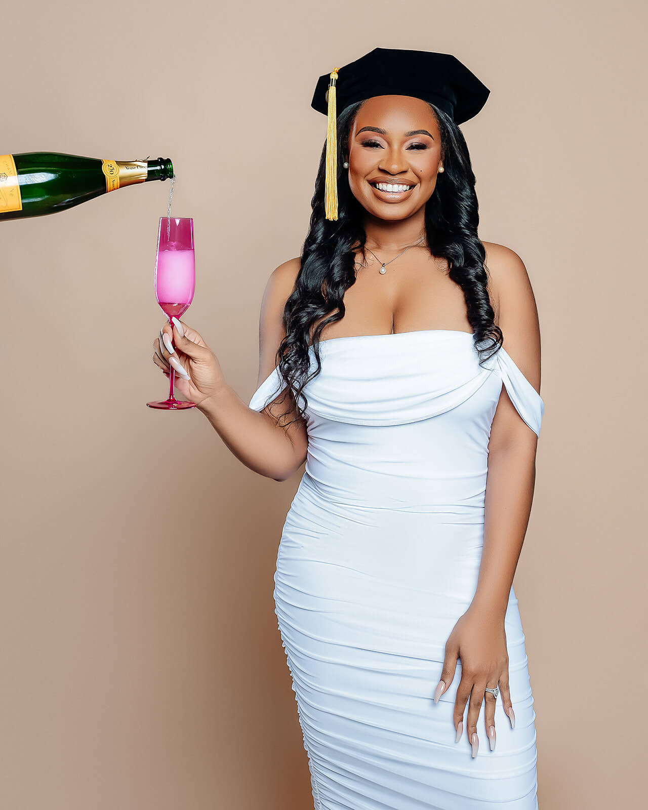 Graduate in cap and white dress receiving champagne pour into pink flute in studio