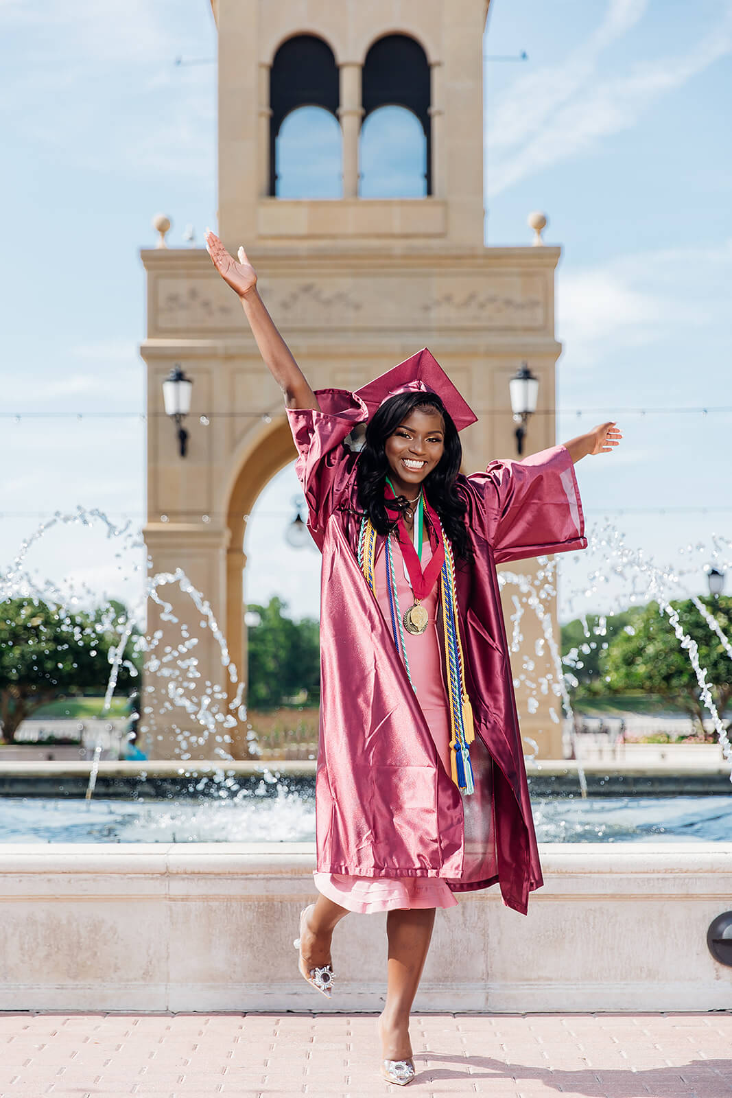 Graduate in maroon cap and gown celebrating with arms raised in front of bell tower fountain