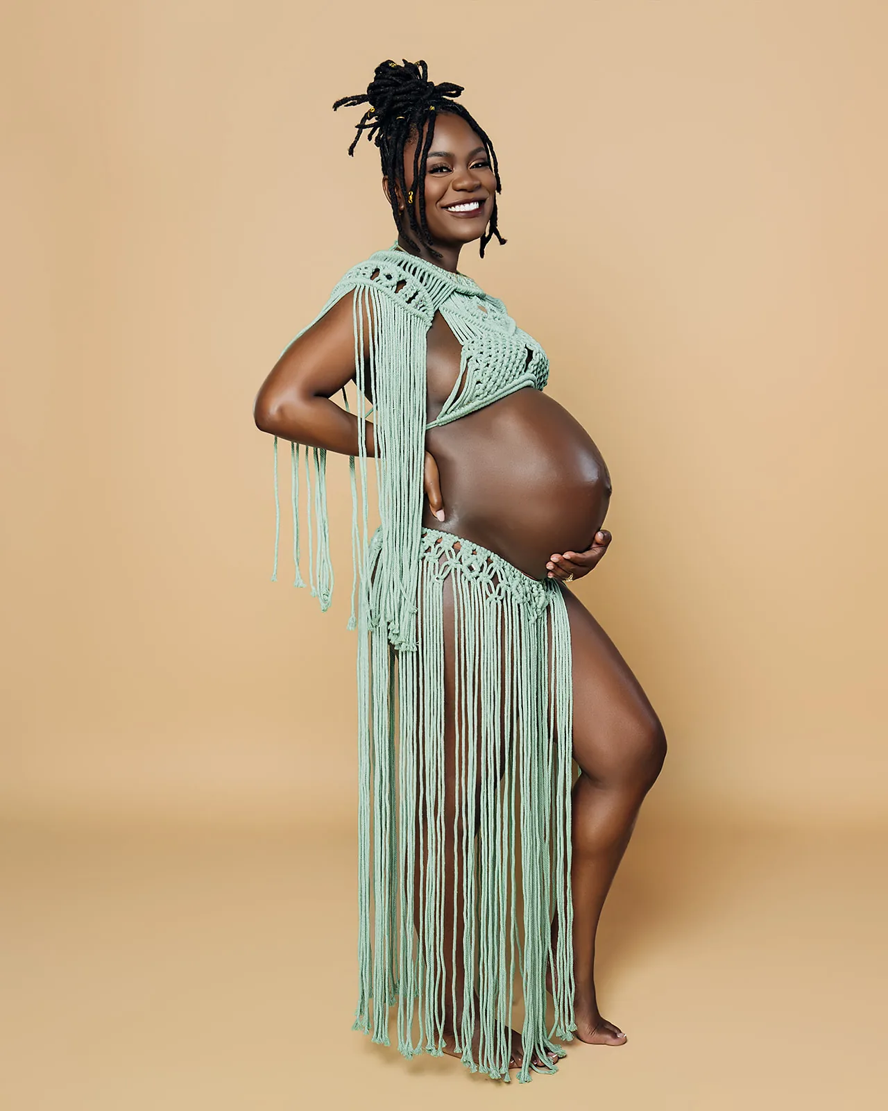 Maternity portrait — expecting mother in green macrame fringe on tan backdrop