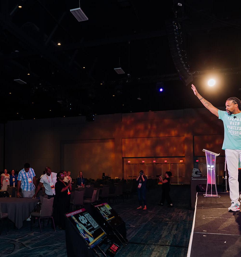 Keynote speaker waving to a large audience from a brightly lit conference stage